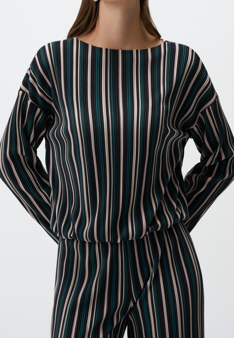 Femme portant un haut à manches longues et un pantalon à rayures verticales noires, blanches, bleu sarcelle et grises, debout les mains derrière le dos.