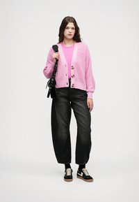 Rosa strikket cardigan med tre store knapper, brukt over en matchende skjorte. Sorte høyhuskede jeans og sorte joggesko med hvite detaljer fullfører stilen.