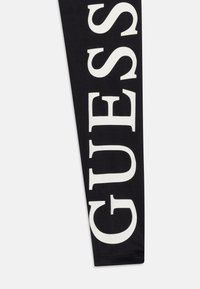 Leggings noirs avec un grand texte blanc en gras écrivant "GUESS." Le tissu est lisse avec une finition mate et une taille standard.
