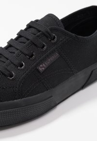 Czarne płócienne sneakersy z fakturowanego materiału, okrągłym noskiem, czarną gumową podeszwą i sznurowadłami w tym samym kolorze. Posiadają małą fioletową naszywkę z logo z boku.