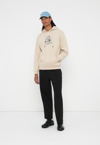 Beige hættetrøje med grafisk design på forsiden, parret med sorte bukser og sorte sneakers. En lyseblå kasket fuldender looket.