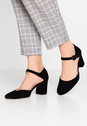 Schwarze Wildleder-Pumps mit abgerundetem Zeh, Knöchelriemen mit einem Knopfdetail und ausgeschnittenen Seiten, kombiniert mit karierten, cropped Hosen.