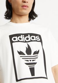White cotton t-shirt featuring a large black adidas logo with a torch design, outlined in a rectangular border. Short sleeves, crew neck.