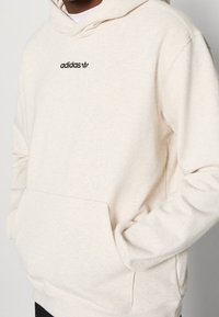 Cremfarbener Kapuzenpullover aus weichem Stoff, mit einer vorderen Tasche und einem schwarzen Adidas-Logo auf der Brust gestickt.