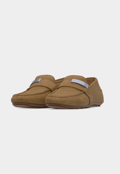 Paire de mocassins en daim beige avec des détails cousus et des accents métalliques argentés sur les sangles, dotés de semelles en caoutchouc antidérapantes.