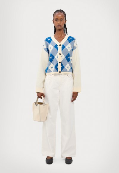 Knitted cardigan in cream with blue argyle pattern; black buttons; paired with white wide-leg pants; holding a small cream handbag.