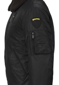 Giacca bomber nera con un colletto marrone imbottito, numerose tasche e un'applicazione testurizzata sulla spalla. Tessuto liscio e resistente.