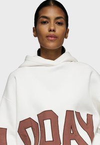 Haut de sweatshirt blanc surdimensionné avec un grand texte graphique marron à l'avant, présentant une texture douce et un design de capuche à double épaisseur.