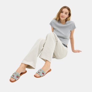 Jonge vrouw zit op de vloer, draagt een lichtgrijs t-shirt, beige broek en grijze slides met kersenborduursel.