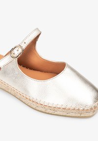 Espadrille plateado metálico con punta redonda y una correa asegurada por una hebilla circular. Presenta bordes cosidos y una parte superior de cuero texturizado.