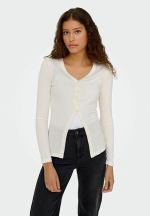 Witte top met lange mouwen en V-hals, met knoopsluiting, voorzien van ribbeltextuur en contrasterende bies. Gecombineerd met donkere spijkerbroeken.