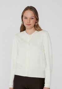Witte cardigan met een ronde halslijn, lange mouwen en knoopsluiting. Zachte textuur en aansluitende stijl. Draag over een donkerbruine broek.