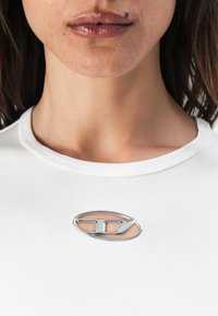T-shirt blanc ajusté avec un col rond et un logo ovale argenté affiché en évidence sur la poitrine, présentant un détail découpé intérieur.