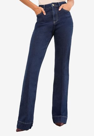 Femeie purtând blugi albastru închis cu talie înaltă și croială dreaptă, cu mâinile în buzunare, asortați cu pantofi maro ascuțiți.