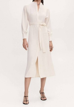 Kvinde iført en cremefarvet midi-kjole med lange ærmer og taljebånd samt sorte højhælede sandaler med stropper, stående foran en ensfarvet baggrund.