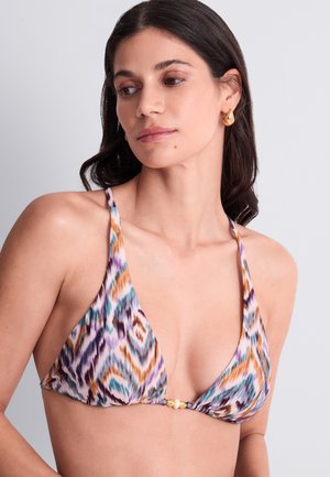Femme aux cheveux foncés portant des boucles d'oreilles en or et un haut de bikini multicolore à motifs avec des bretelles fines et un détail en perles au centre.