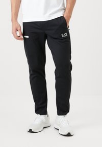 Sötétkék sportnadrág keskeny szabással, sima anyagból készült. Jellemzői közé tartozik a cipzáras zsebek és egy kis logó a felső combon.