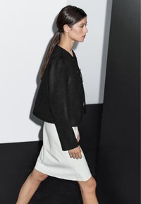 Kvinna med mörkt hår i hästsvans går iförd en svart långärmad jacka och en vit knälång kjol mot en svart-och-vit bakgrund.