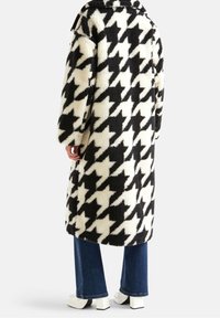 Płaszcz w czarno-biały wzór pepitki, wykonany z puszystej tkaniny, sięgający do kolan, z szerokim kołnierzem i oversize'owymi rękawami. W zestawieniu z niebieskimi dżinsami i białymi botkami.