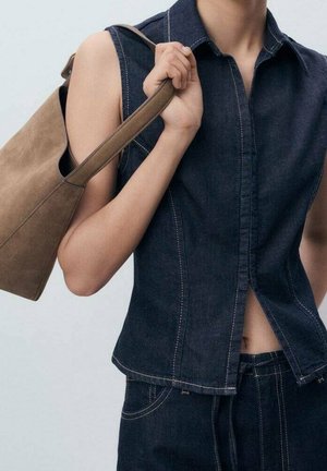 Persona con top vaquero sin mangas de color azul oscuro y jeans, sosteniendo un bolso marrón de ante en el hombro derecho.