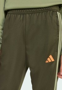 Tmavě zelené sportovní kalhoty s texturovaným povrchem, s elastickým pasem, světlými pruhy a oranžovým logem na boku.