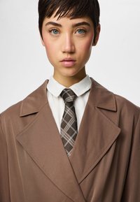 Νεαρό άτομο με κοντά σκούρα μαλλιά φοράει καφέ παλτό, λευκό πουκάμισο με γιακά και καφέ γραβάτα με καρό μοτίβο, σε απλό λευκό φόντο.