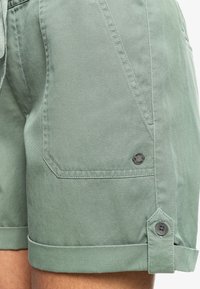 Gros plan sur la poche d'un short en denim vert clair avec deux boutons noirs et un ourlet roulé sur la cuisse d'une personne.