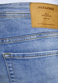 Vaqueros de mezclilla azul claro con una textura suave, que presentan un parche de cuero etiquetado "JACK & JONES" y costuras amarillas a lo largo de las costuras.
