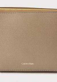 Τσεπάκι από δέρμα σε απόχρωση ταουπ με χρυστό φερμουάρ και το λογότυπο «Calvin Klein» χαραγμένο σε χρυσό κοντά στο κέντρο του κάτω μέρους.