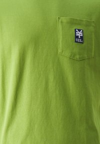 Zelena bombažna majica z žepom na prsih in modrim vezenim logotipom, gladka tekstura in klasičen kroj z okroglo ovratnikom.