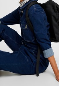 Personne portant une combinaison en jean bleu foncé et un col roulé gris, assise avec un sac à dos noir et les manches retroussées visibles.