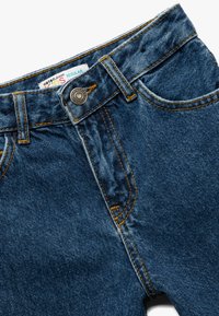 Jeans de mezclilla en azul oscuro con costuras naranjas, que presentan un bolsillo frontal, cierre de botón metálico y una etiqueta que dice "Katrin Jeans Regular."