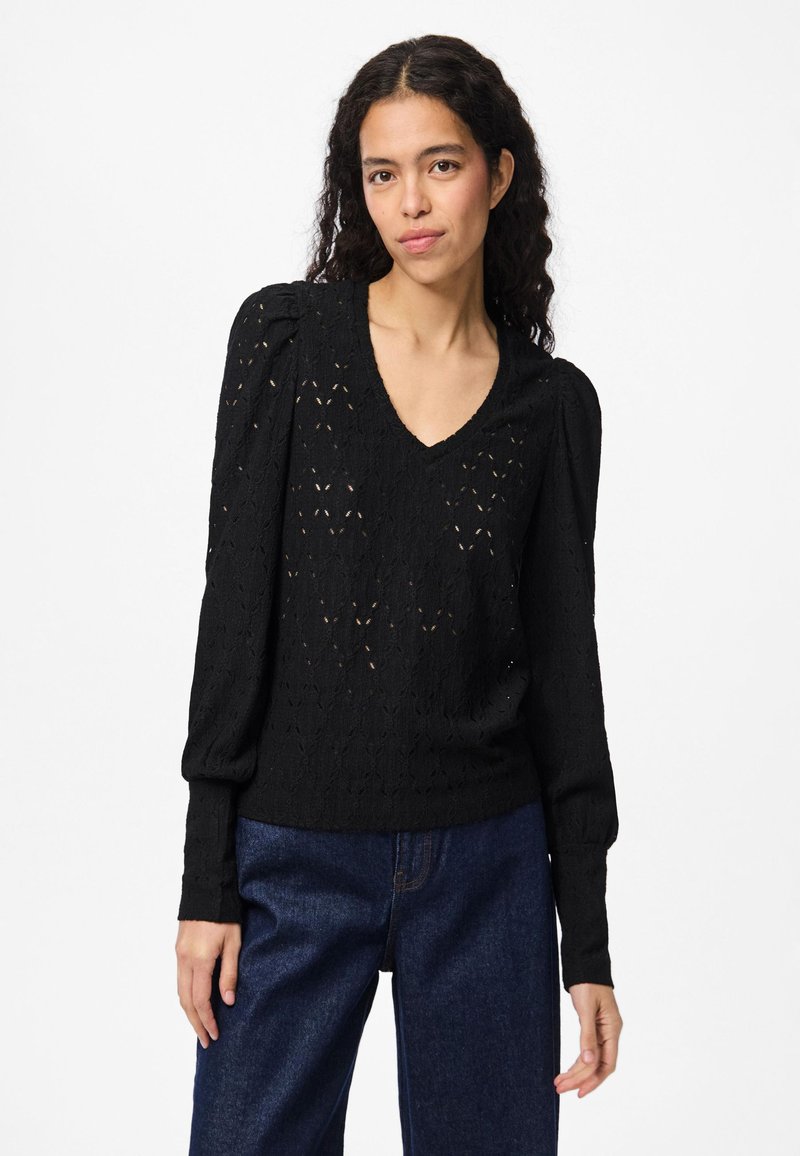 Svart genser med lange ermer og V-hals, med teksturert åpent strikkedesign og lett puffede skuldre. Kombinert med denimbukser.