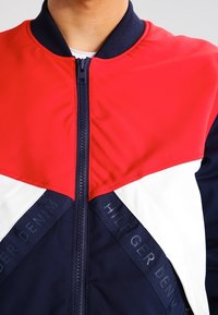 Chaqueta bomber roja y azul marino con acentos blancos, que presenta un cierre frontal, cuello acanalado y la marca "HILFIGER DENIM" en los laterales.