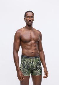 Fekete háttérrel rendelkező, zöld trópusi levélmintás boxer rövidnadrág, fekete elasztikus derékpánttal, amelyen a "BJÖRN BORG" felirat szerepel.
