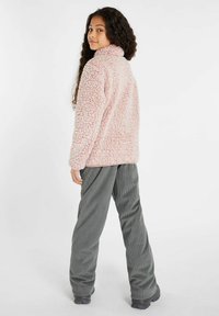 Fleecejacke in sanftem Rosa mit hohem Kragen und strukturierter, flauschiger Oberfläche, kombiniert mit lockeren grauen Cordhosen und schwarzem Schuhwerk.