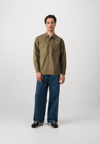 Olive green, button-up shirt with two front pockets, paired with blue wide-leg pants and black sneakers with white accents.