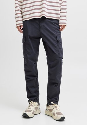 Personne portant un pantalon cargo noir, des baskets beige et blanches, et une chemise à manches longues beige et blanche rayée.