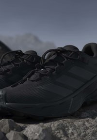 Chaussures de sport noires avec semelles texturées et lacets reposant sur un sol rugueux et rocheux sous un ciel gris.