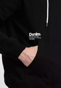 Czarna bluza z kapturem wykonana z bawełny o teksturowanym wykończeniu, z kapturem ze sznurkiem i białym logo po lewej stronie z detalami produktu.