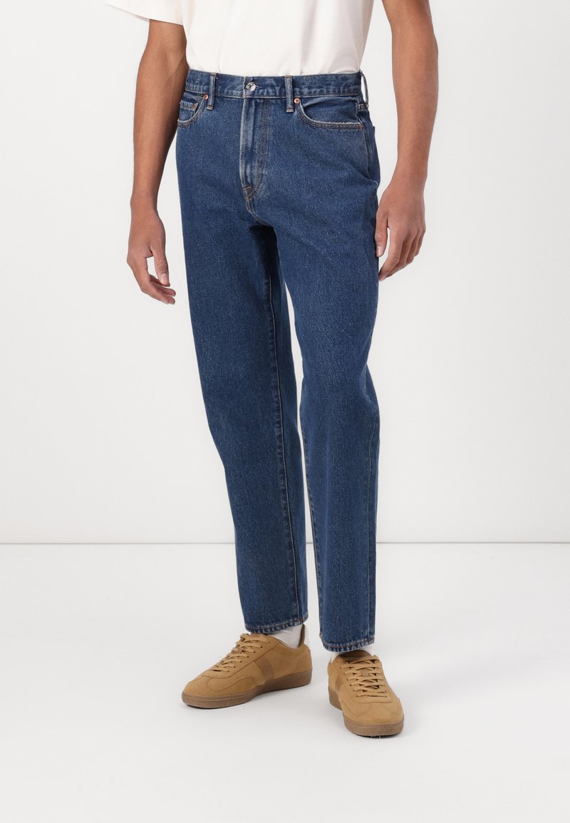 Temno modri jeans z visokim pasom, ravnimi hlačnicami in subtilnimi zbledelimi podrobnostmi. V kombinaciji z bež semiš športnimi čevlji.