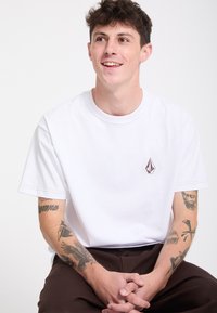 Jeune homme aux cheveux courts et bouclés souriant, portant un t-shirt blanc et un pantalon marron, avec plusieurs tatouages sur les deux avant-bras.