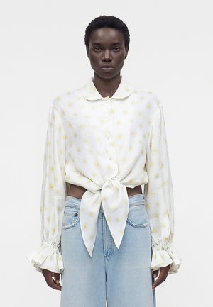 Nő fehér, gombos blúzt visel, amelyen sárga margaréta mintázat van, a deréknál megkötve, világoskék, magasderekú farmerrel párosítva.