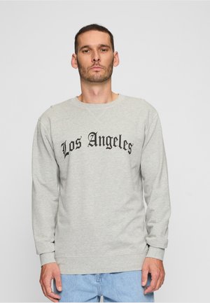 Mann mit kurzen dunklen Haaren und Bart, trägt einen hellgrauen Pullover mit der Aufschrift „Los Angeles“ und hellblaue Jeans, steht vor einfachem Hintergrund.