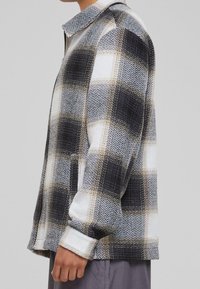 Mężczyzna w czarno-biało-szarym koszuli w kratę z długimi rękawami i kołnierzykiem, stojący na jednolitym szarym tle.