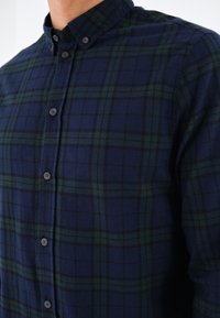 Chemise à carreaux bleu marine et vert foncé, boutonnée avec col, sur la partie supérieure du torse d’une personne sur un fond uni.