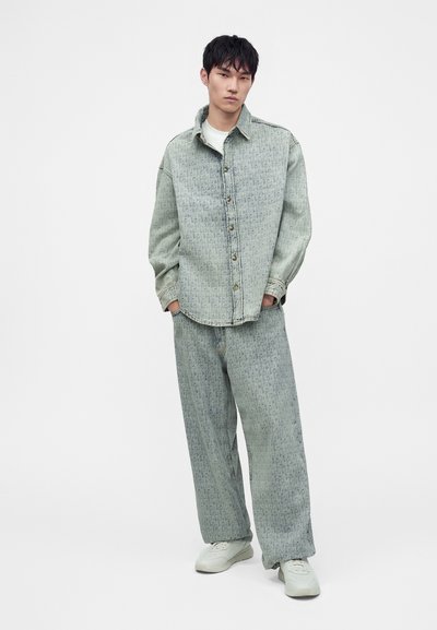 Νέος άνδρας στέκεται με τα χέρια στις τσέπες, φοράει ένα ανοιχτοπράσινο πουκάμισο ντένιμ με σχέδιο και αντίστοιχο φαρδύ παντελόνι με λευκά αθλητικά παπούτσια.