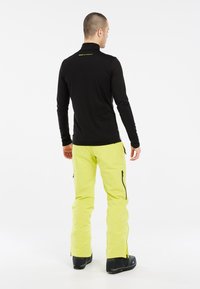 Zwarte langermige shirt met een hoge kraag, gecombineerd met fluorescent gele skibroek met zijritsen en zwarte laarzen.