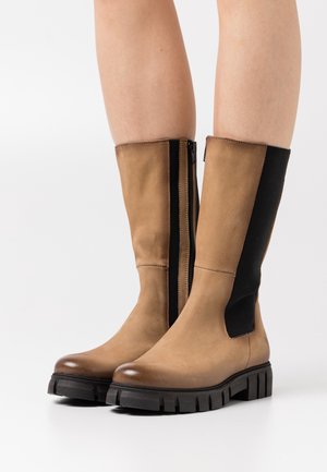 Botas de cuero marrón hasta la rodilla con un panel lateral elástico negro, suela de goma con relieve y un zipper lateral para facilitar su uso.