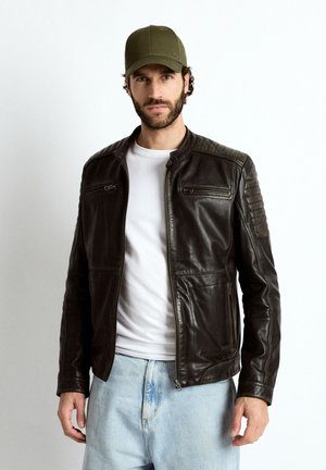 Mand iført olivengrøn cap, sort læderjakke med lynlåslommer, hvid T-shirt og lyseblå jeans, stående mod en ensfarvet baggrund.